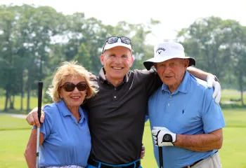 Three older adults stand arm in arm on a golf course.