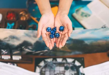 two hands holding dice with dungeons and dragons game items in background