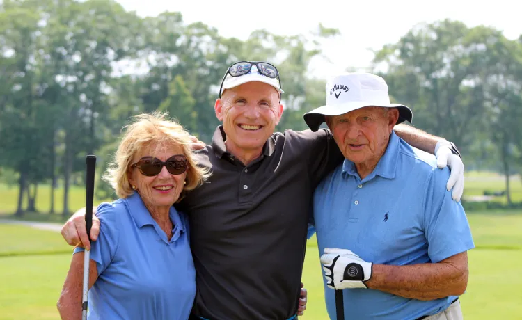 Three older adults stand arm in arm on a golf course.