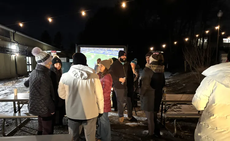 group of adults outside with string lights and large screen of football