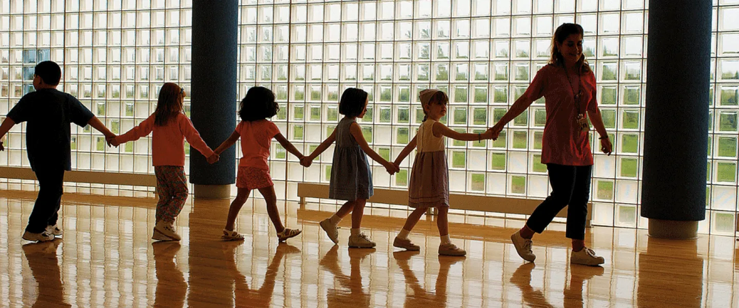 A teacher leads a line of children holding hands in a gymnasium