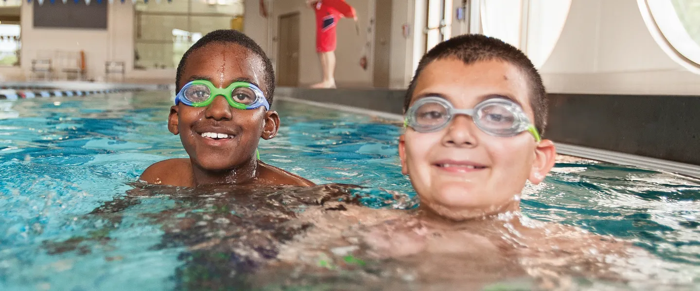 Two boys wearing swim goggles smile while treading water in an indoor pool