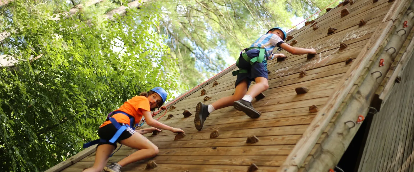Two kids secured with harnesses and ropes climb up a wooden climbing tower
