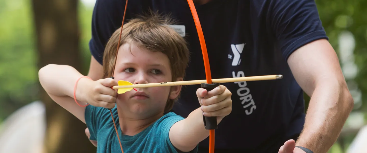 A boy holds a bow and arrow ready to shoot while an archery instructor stands behind him