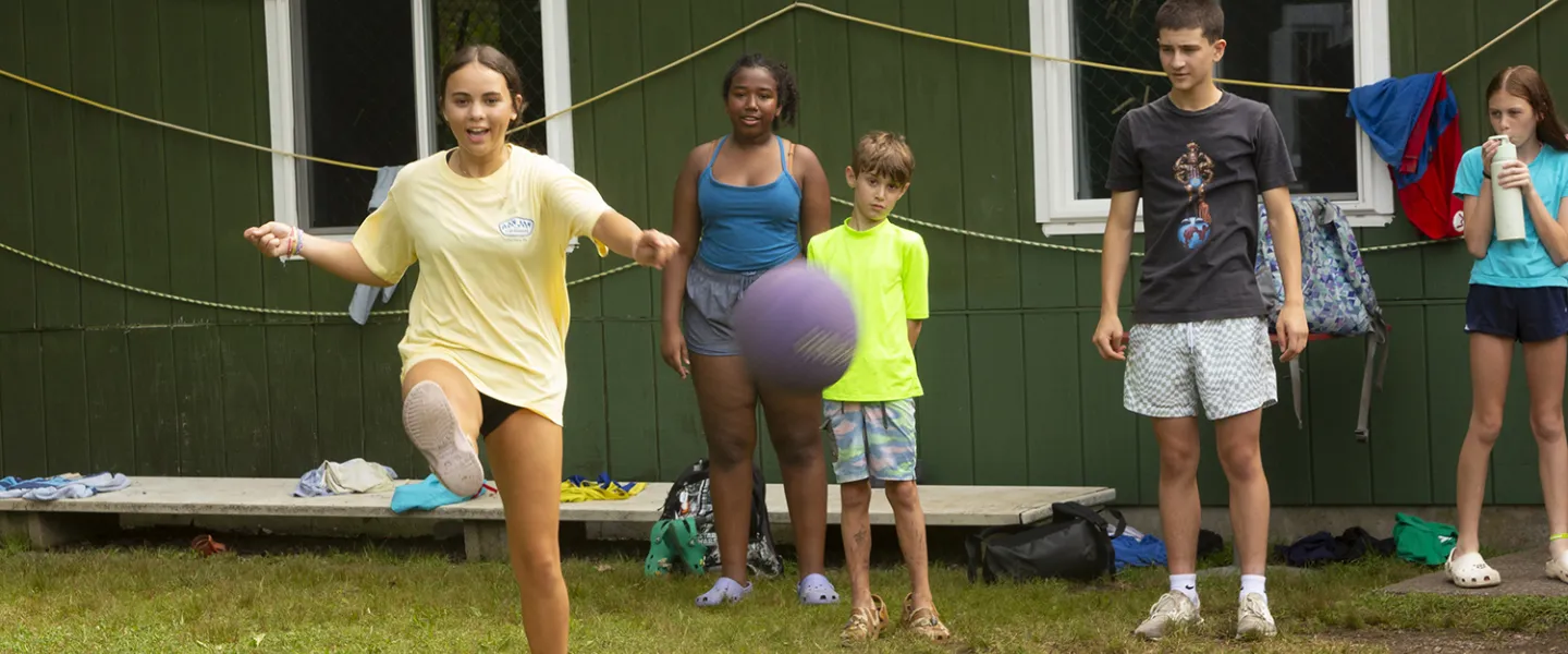 Campers gathered around playing kickball, as one girl takes a big kick.