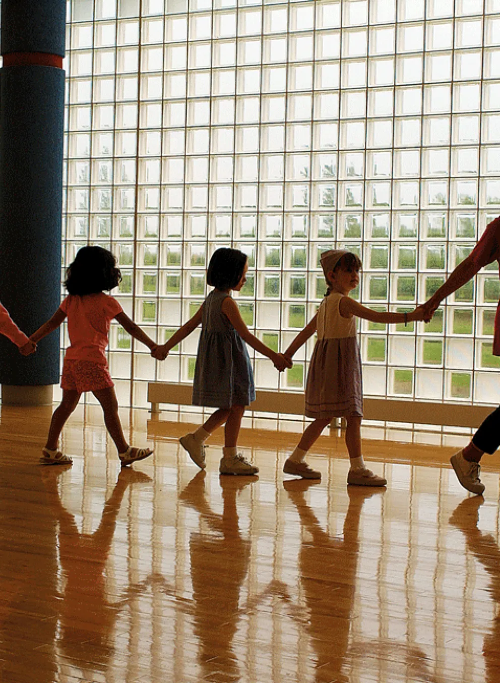 A teacher leads a line of children holding hands in a gymnasium