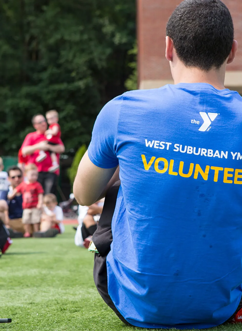 The back of a man wearing a shirts that reads "volunteer" is shown in the foreground while children sit in the background