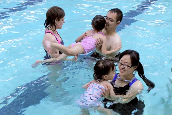 A swim instructor talks to two parents who are each holding a child in an indoor swimming pool