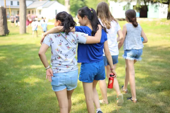 Two girls walking across a grassy field with their arms draped over each other