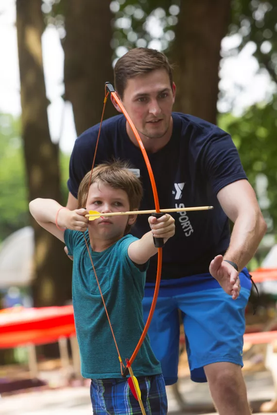 A boy holds a bow and arrow ready to shoot while an archery instructor stands behind him