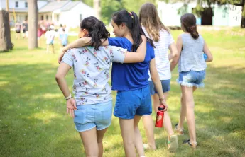 Two girls walking across a grassy field with their arms draped over each other