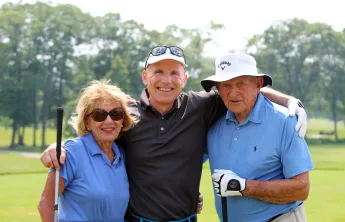 Three older adults stand arm in arm on a golf course.