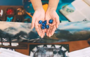 two hands holding dice with dungeons and dragons game items in background