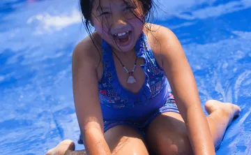 A young asian girl is laughs and smiles while she sits at the end of a slip and slide soaking wet
