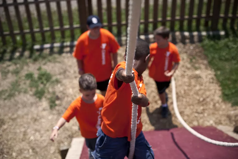 A camper at the YMCA climbs a rope while other campers cheer him on.