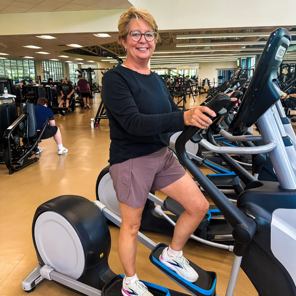 A woman stands on an elliptical, smiling at the camera.
