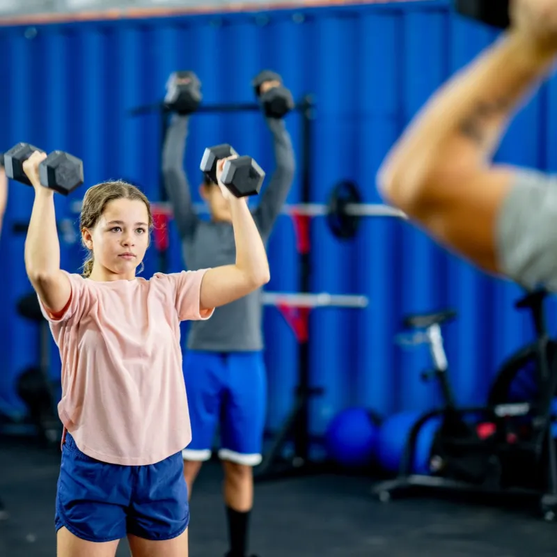 A child lifts two dumbells above their head