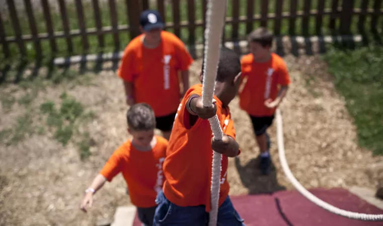 A camper at the YMCA climbs a rope while other campers cheer him on.