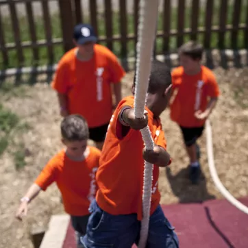 A camper at the YMCA climbs a rope while other campers cheer him on.