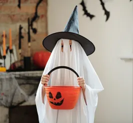 a child celebrating halloween. they are dressed as a ghost with a witch's hat and holding an orange trick-or-treat bucket