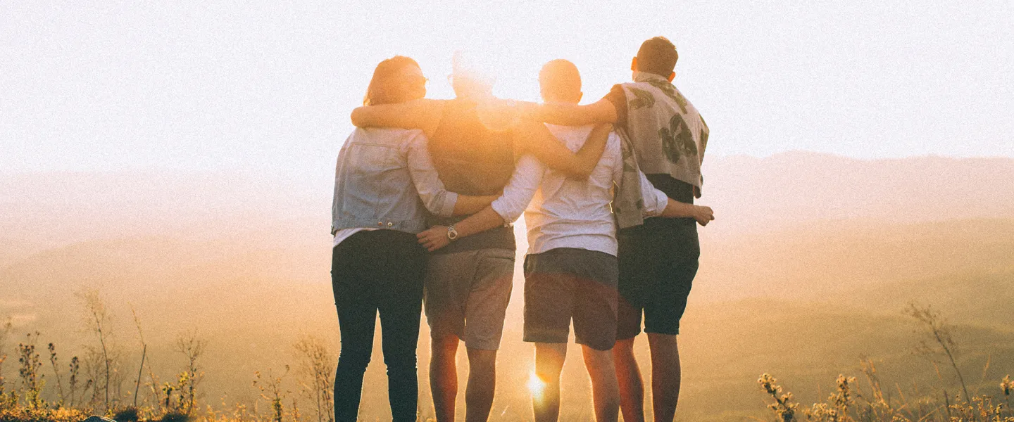 the backs of four adults with their arms around one another. they are looking at a sun setting over low mountains