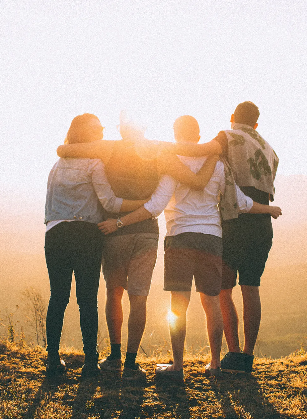 the backs of four adults with their arms around one another. they are looking at a sun setting over low mountains