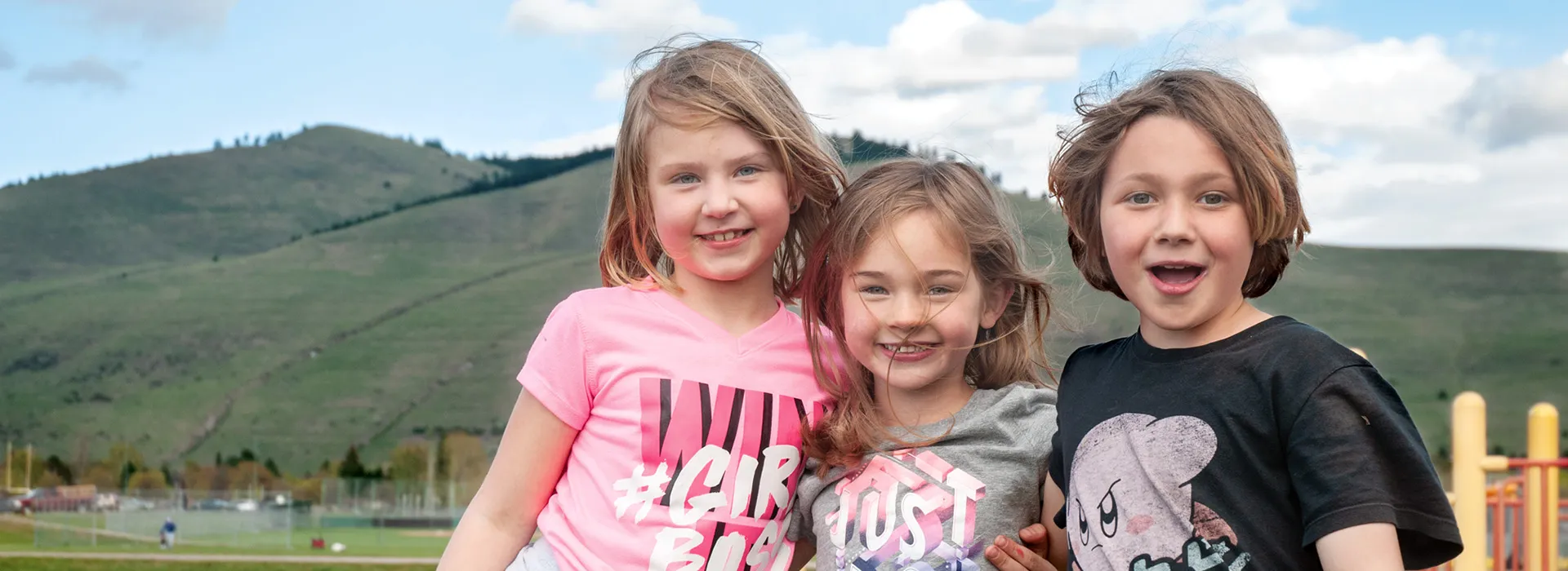 three smiling children standing in front of a large hill with their hair blowing in the wind. 