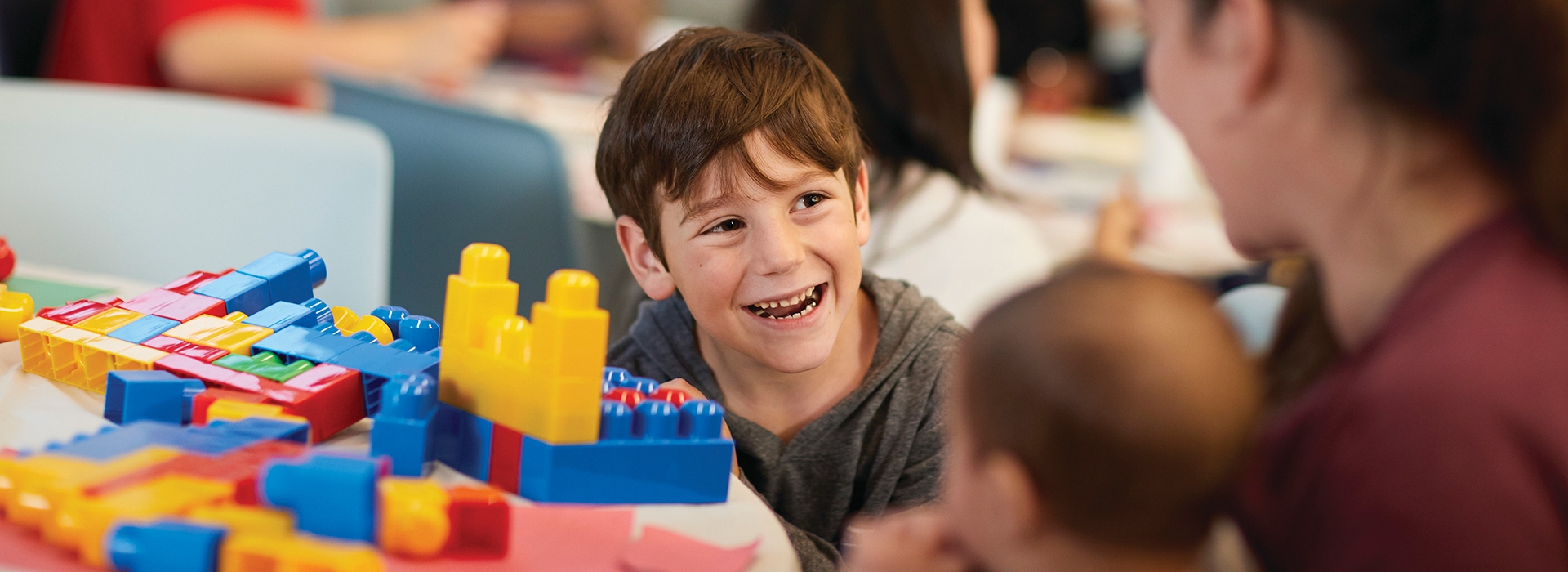 child laughing at table with legos. there is a parent and baby in the foreground