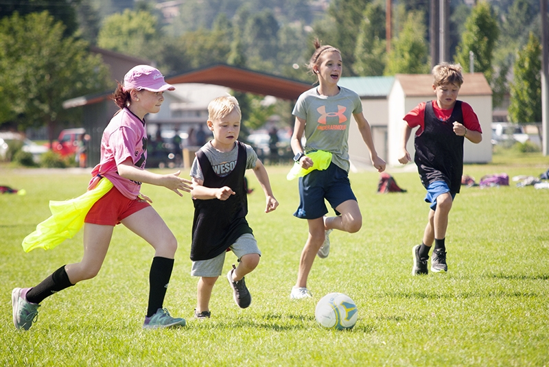 four children of various ages play soccer outdoors during summer camp