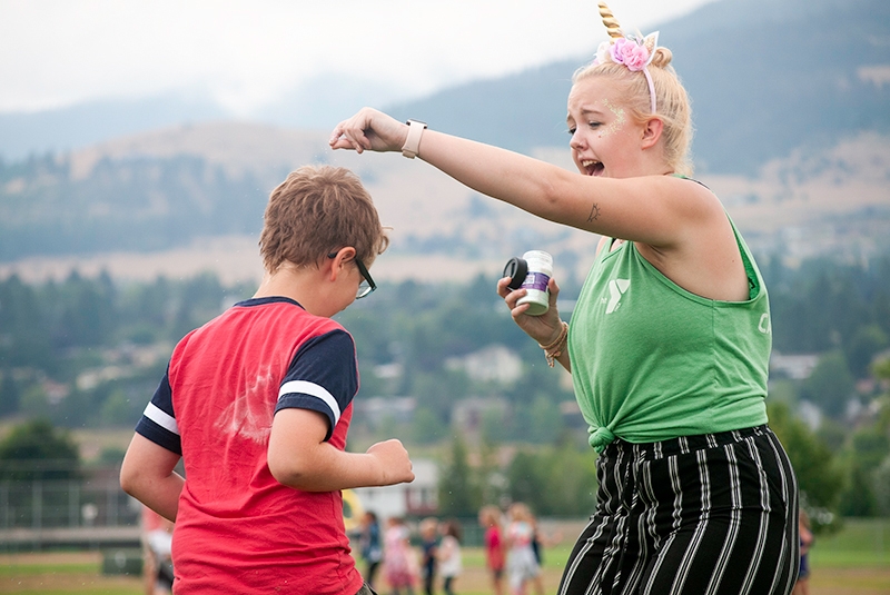 camp counselor with fake unicorn horn sprinkling glitter on the head of a camper with glasses
