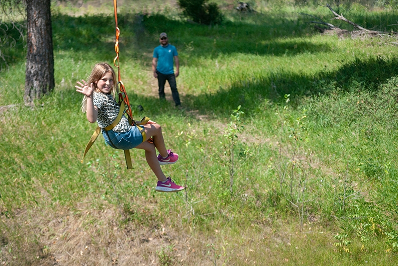 child with long hair waving and riding a zipline in the forest with a counselor in the background
