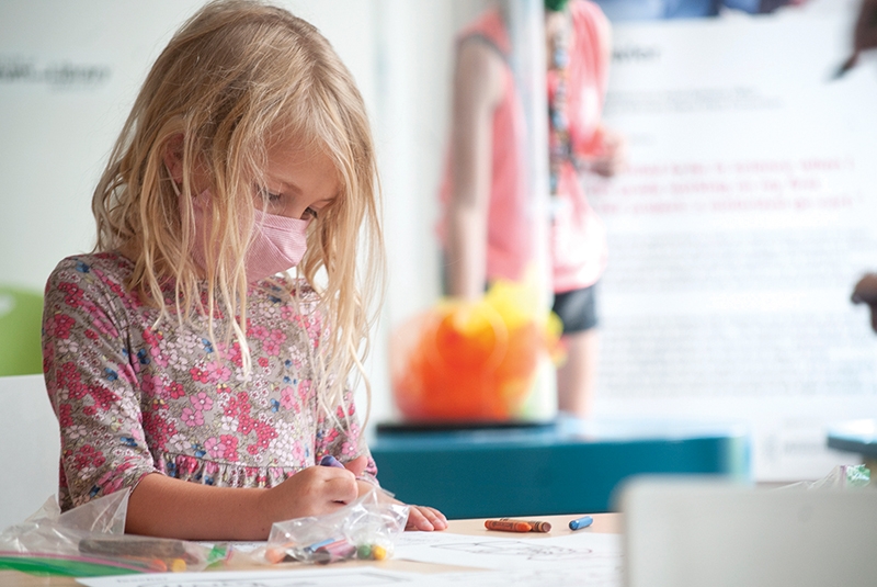 a young child with shoulder-length blonde hair and a pink dress is wearing a mask and coloring at a table