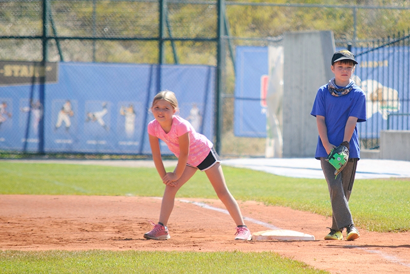 child in pink shirt on first base preparing to run to second base with another child in baseball hat guarding them