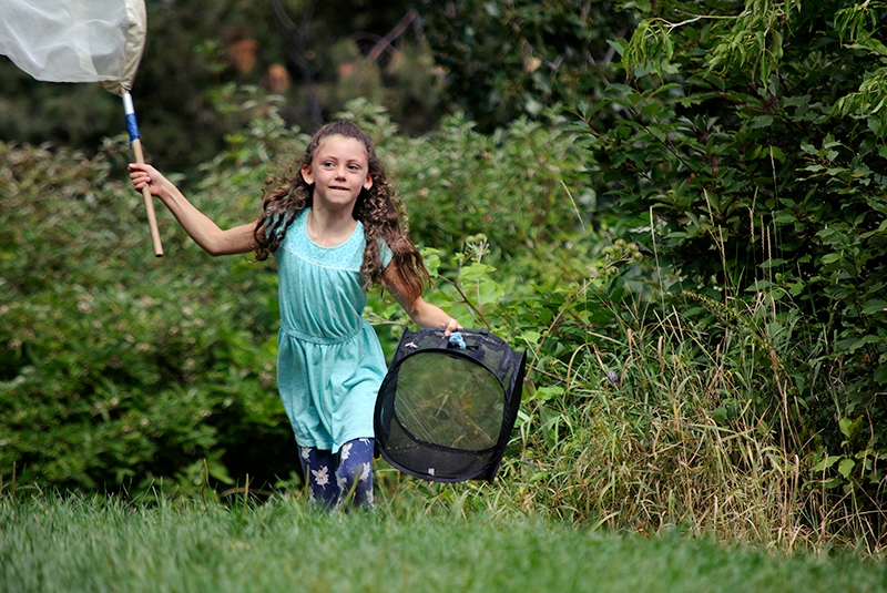 young child with long curly hair and teal dress running outdoors with a bug net and a bug catcher