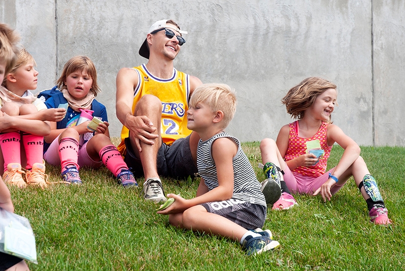 counselor in yellow basketball jersey, baseball hat, and sunglasses sitting in the grass and laughing with a group of young campers