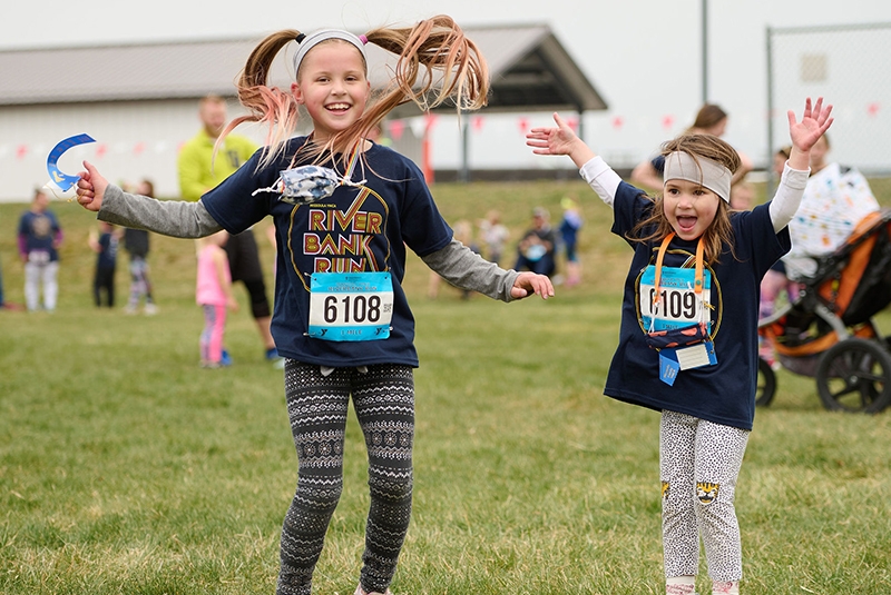 two young children are having fun and jumping. they are wearing race bibs and shirts that say riverbank run. they are preparing to run a 1 mile race.