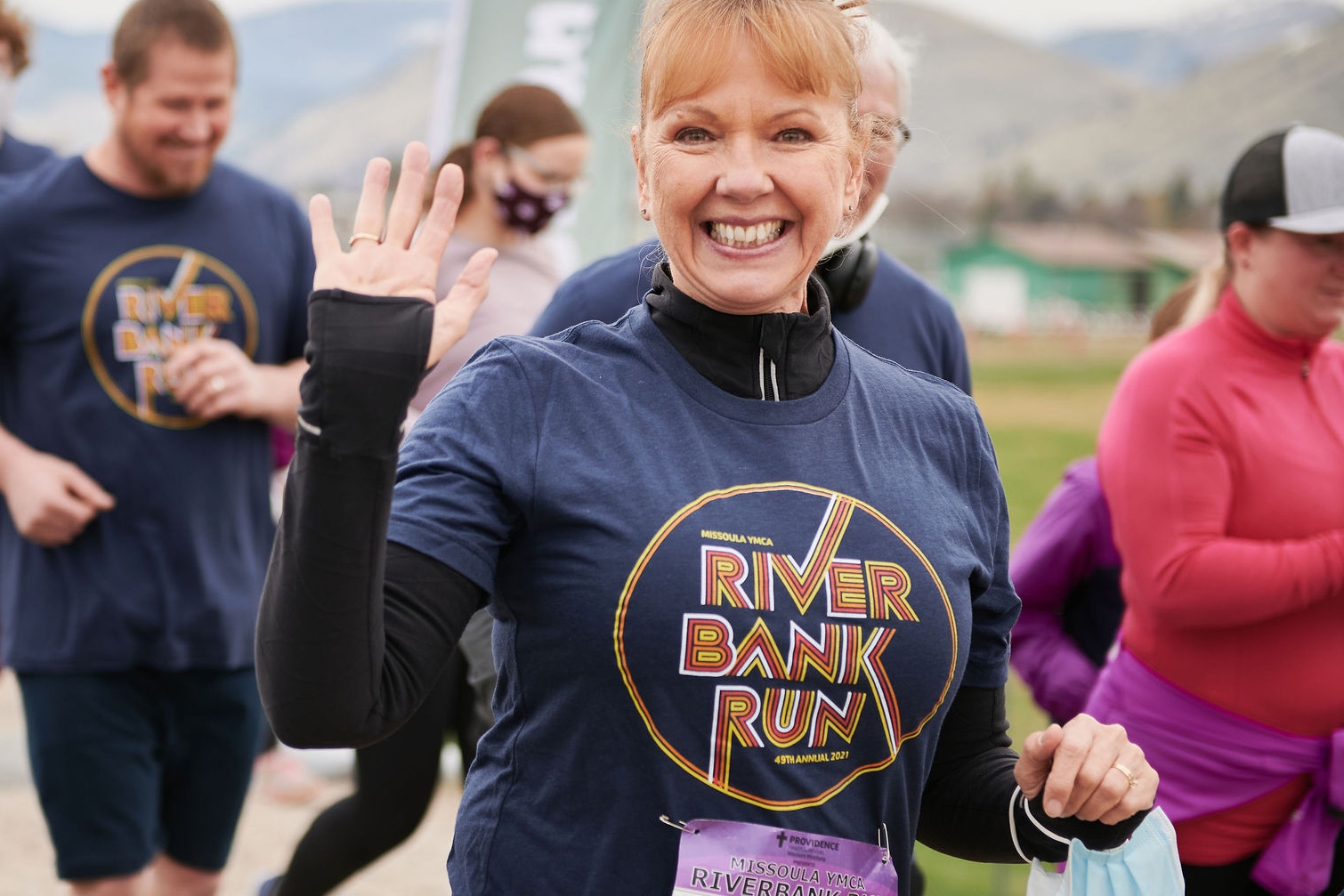 a woman smiling and waving at a camera while running a race and wearing a shirt that says riverbank run