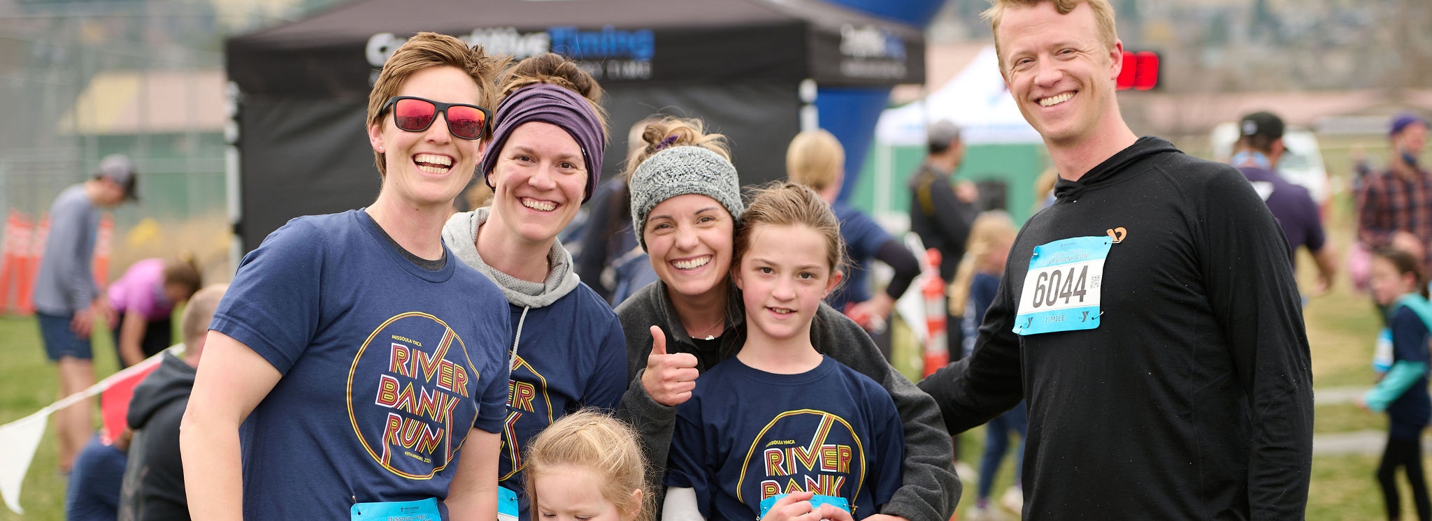 family of six wearing ymca riverbank run shirts with race bibs and smiling at the camera. they have just finished a running race.