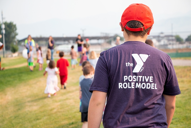 a young adult walks away from the camera. he is wearing a purple shirt with the ymca logo. his shirt says positive role model. he is also wearing a red baseball hat. in front of him is a group of small children, also with their backs to the camera.