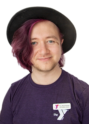 headshot of a smiling adult with purple hair and a black felt hat 