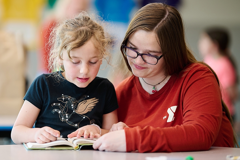 ymca counselor with glasses and a red shirt helps a young child read a book