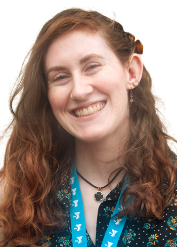 headshot of a smiling woman with long brown-red curly hair. she has dangly earring, a teal lanyard, and a black blouse with flowers. 