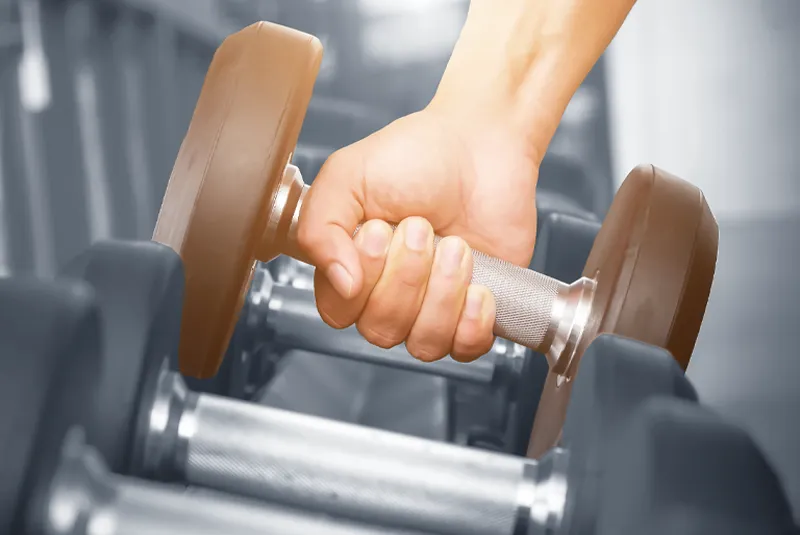 Close-up of a person's hand gripping a dumbbell in a gym setting.