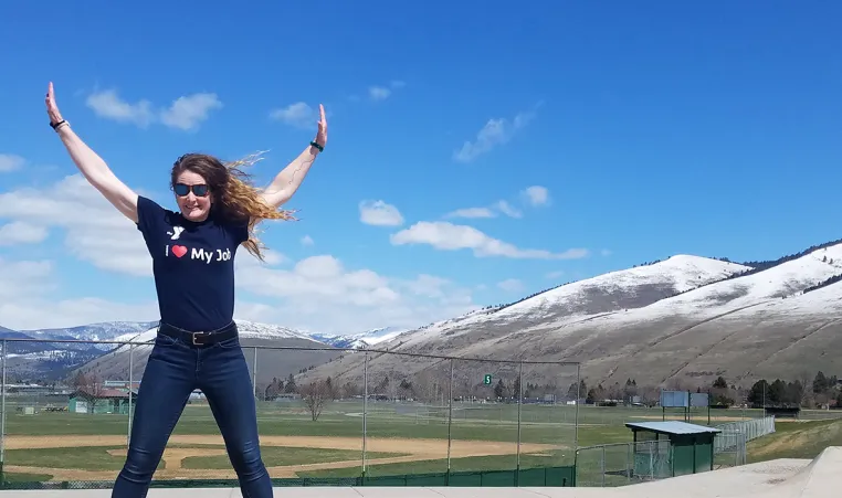 young woman with brown hair wearing a black shirt that says i love my job. she has sunglasses and is jumping in a city park. there are blue skies and snowy mountains in the background.