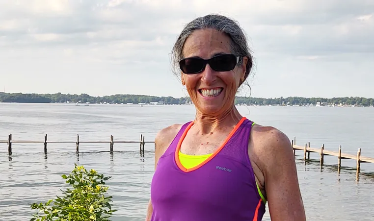 an adult woman with a purple tank top and sunglasses with her grey hair pulled back. she is smiling and standing in front of a big lake 