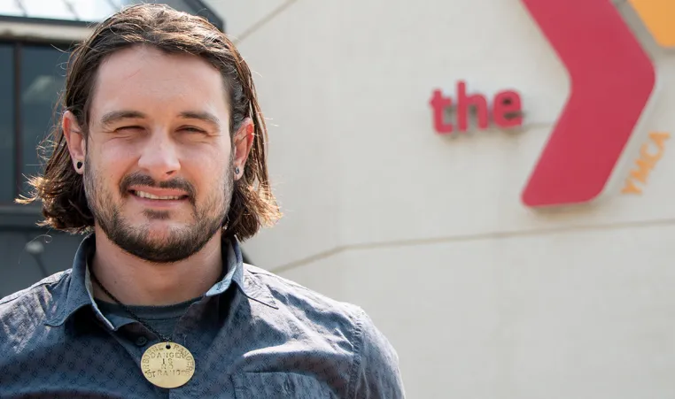 an adult man with brown shoulder-length hair in front of a ymca and squinting into the sun. he wears a circular gold necklace honoring an army ranger.