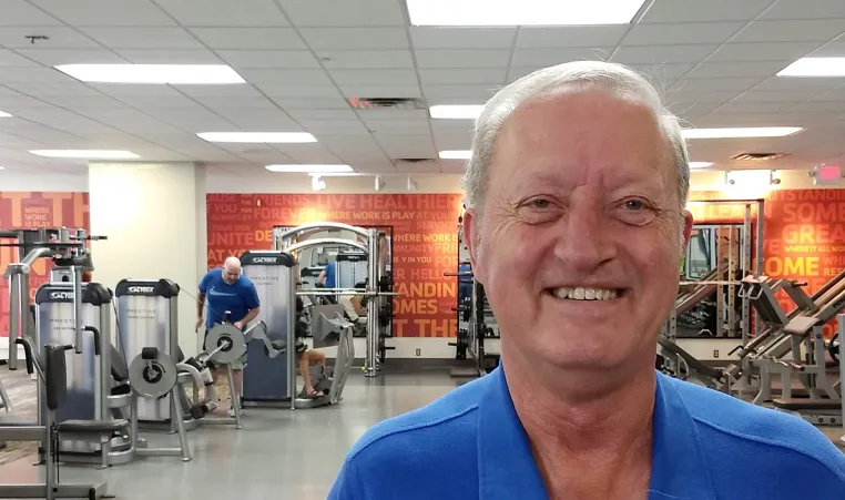 a man with white hair and a blue shirt stands in a busy fitness gym