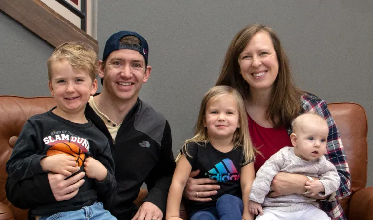 two parents sit with their three young children on brown leather chairs in front of a grey wall