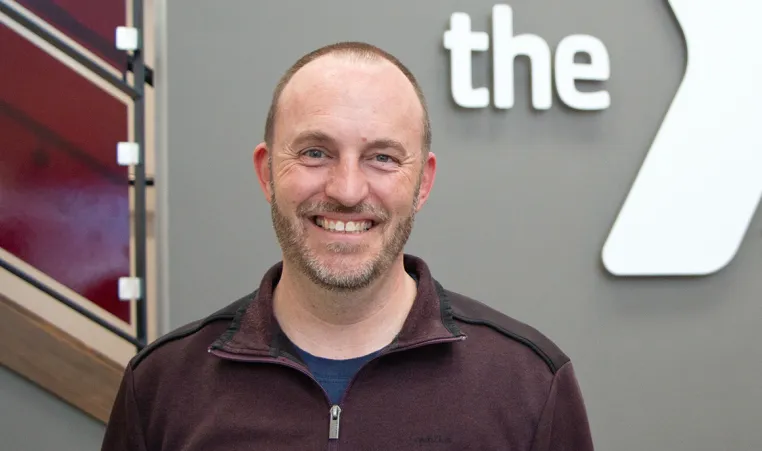 a photo of a man in a maroon zip jacket and light beard stubble smiles at the camera