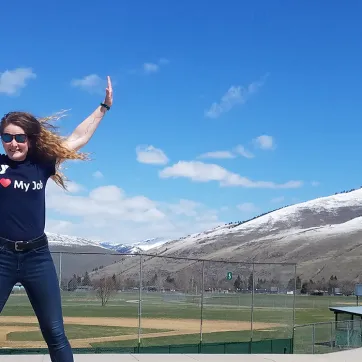 young woman with brown hair wearing a black shirt that says i love my job. she has sunglasses and is jumping in a city park. there are blue skies and snowy mountains in the background.